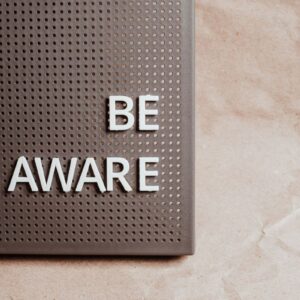A black pinboard on a light background with letters pinned in spelling out BE AWARE