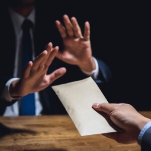 A dark image showing a hand holding a sealed envelope aloft across a wooden table at someone sat opposite holding their hands up in a way that seems to be saying no to illustrate a bribery act
