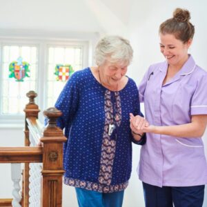 A smiling care worker helping an elderly lady at the top of some stairs with stain glassed windows behind them