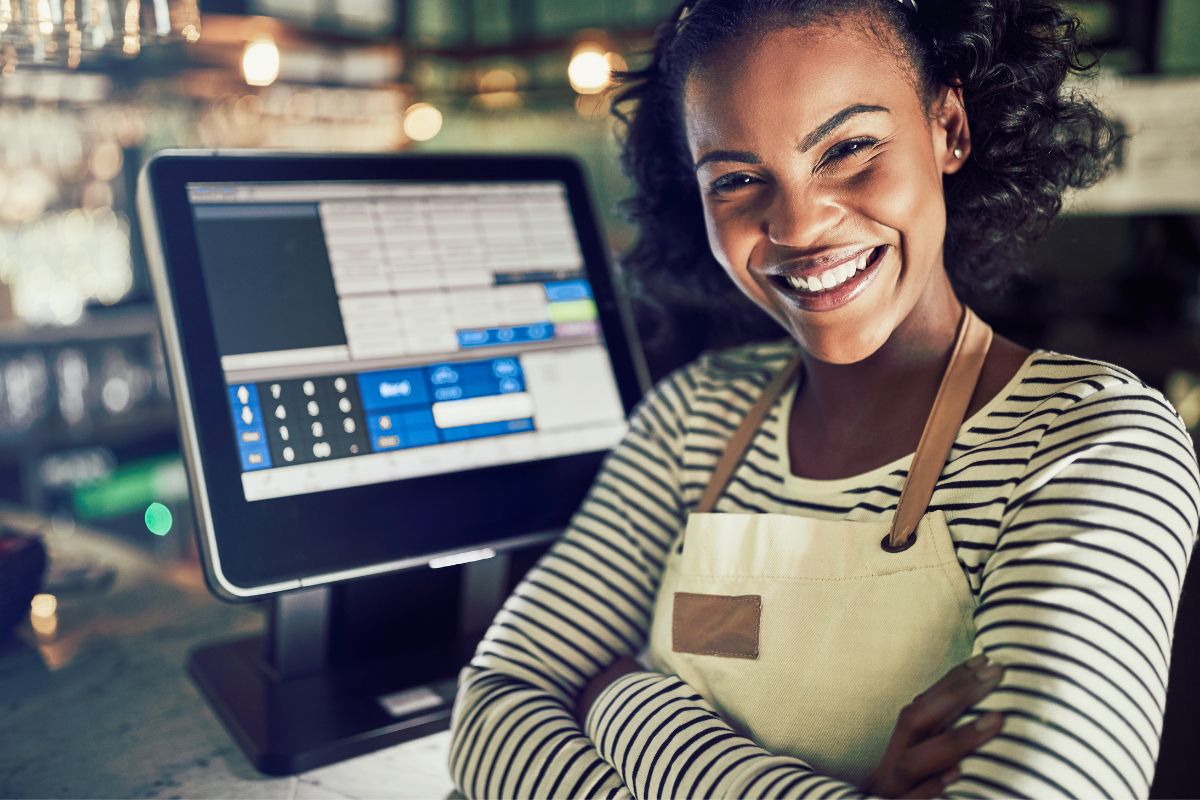 A smiling worker with apron on and arms folded in front of a till screen