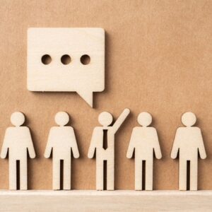 A row of seven wooden 'people' all looking the same apart from the middle one which has a tie on and hand raised and a speech bubble above them like they are asking a question and speaking