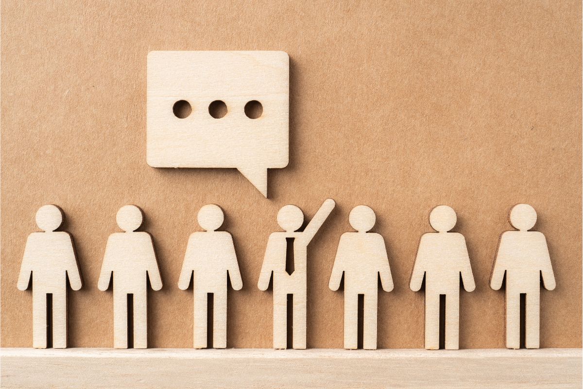 A row of seven wooden 'people' all looking the same apart from the middle one which has a tie on and hand raised and a speech bubble above them like they are asking a question and speaking