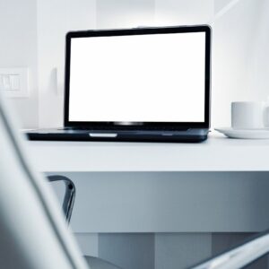 A laptop displaying a white screen on w white table with white walls and a white cup and saucer and light beside it