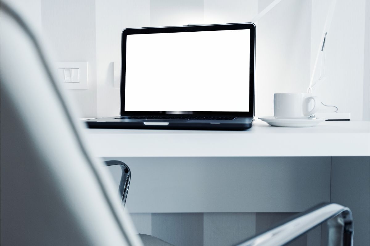 A laptop displaying a white screen on w white table with white walls and a white cup and saucer and light beside it