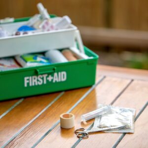 A green First Aid box filled with first products and a bandage scissors, syringe and plaster roll in front on a table top