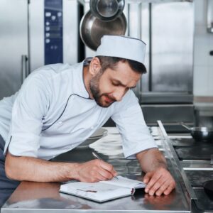 A man in chefs whites in a commercial kitchen resting on a stainless steel counter writing in a notepad