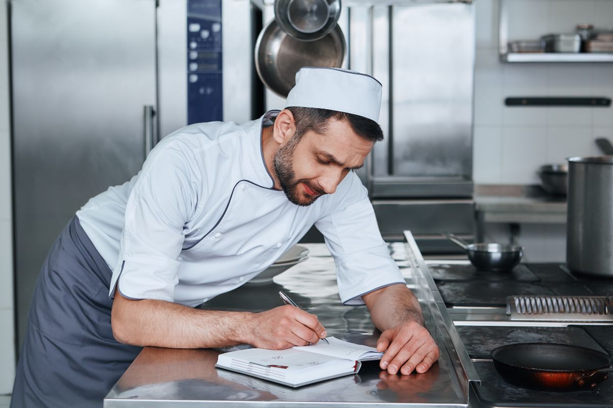 A man in chefs whites in a commercial kitchen resting on a stainless steel counter writing in a notepad
