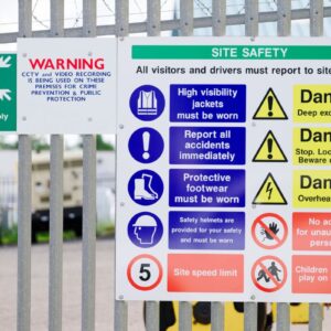 A railing with Health and Safety signs on display, one is a fire assembly point, one a WARNING that CCTV and video recording is in use and the final a large Site Safety sign.
