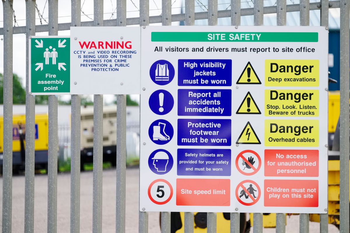 A railing with Health and Safety signs on display, one is a fire assembly point, one a WARNING that CCTV and video recording is in use and the final a large Site Safety sign.
