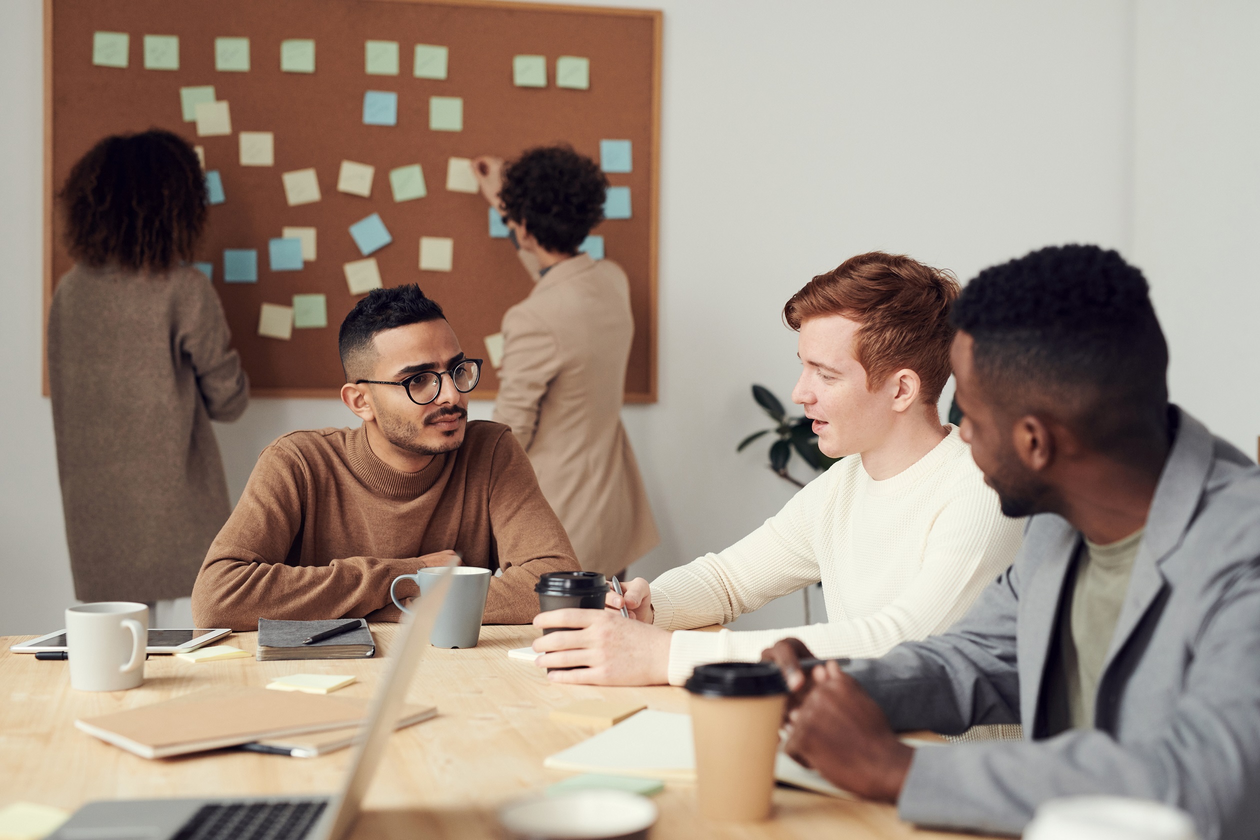 Image showing an Office Meeting with three sat at a table discussing matters with mugs and coffee cups and two stood at a pinboard behind placing more post-it notes on it