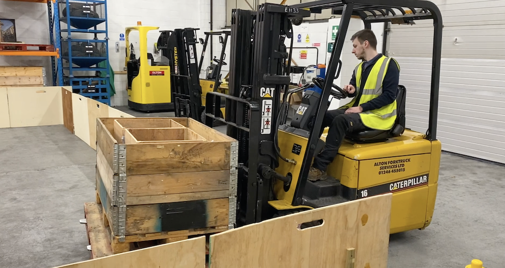 Image within a factory or warehouse setting showing a man sat in an high viz vest operating a forklift and moving a load of wooden crates