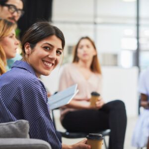 Image to illustrate team working with five colleagues sat around some with takeaway coffee cups and some with notepads and notes. There front lady smiling directly at the camera.