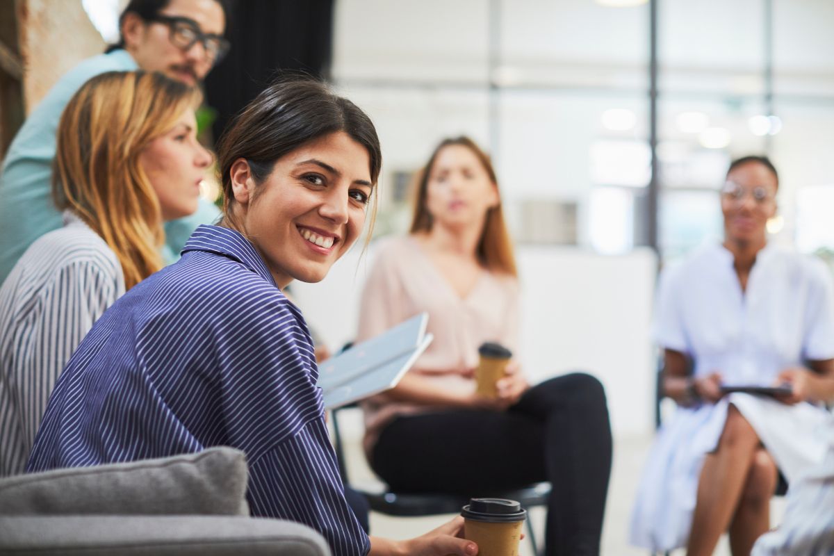 Image to illustrate team working with five colleagues sat around some with takeaway coffee cups and some with notepads and notes. There front lady smiling directly at the camera.